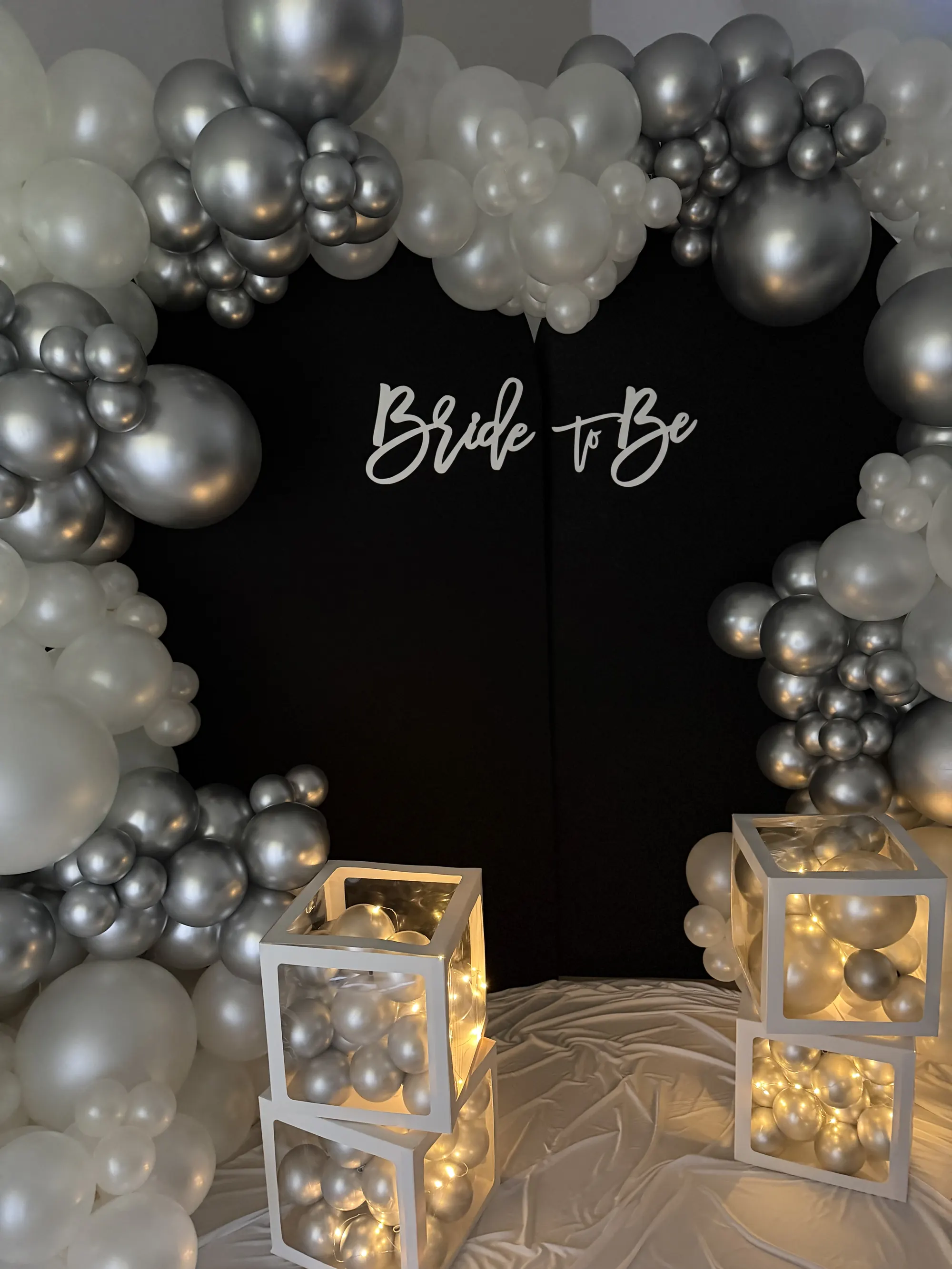 A silver and pearl balloon garland framing a black Bride to Be backdrop, with glowing balloon boxes